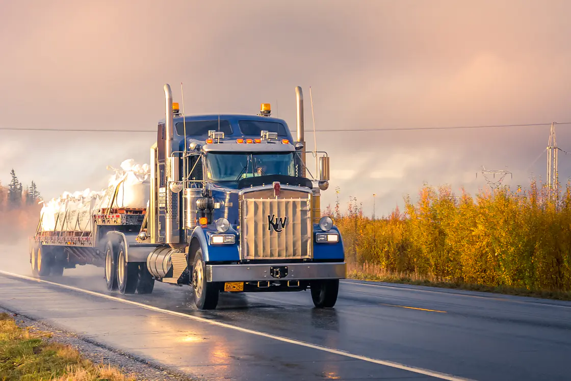 A blue tractor pulling a flatbed load on a wet highway at sunrise