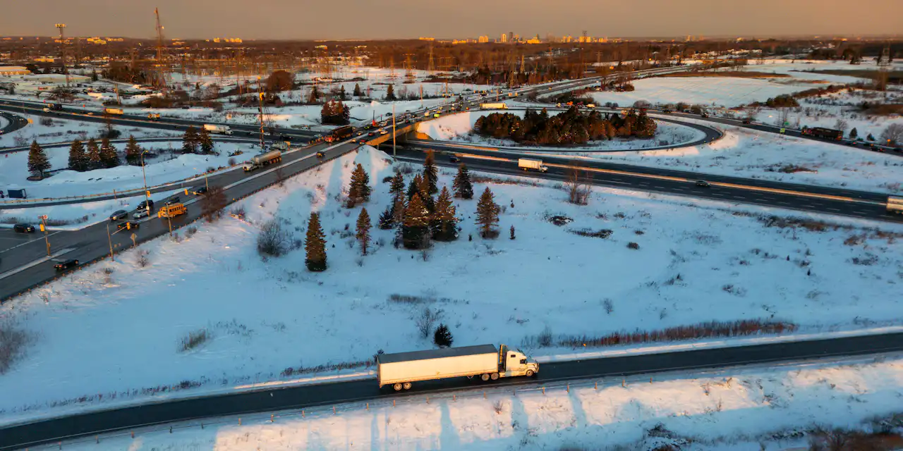 A tractor-trailer on a snow-lined highway beside a large interchange at sunset